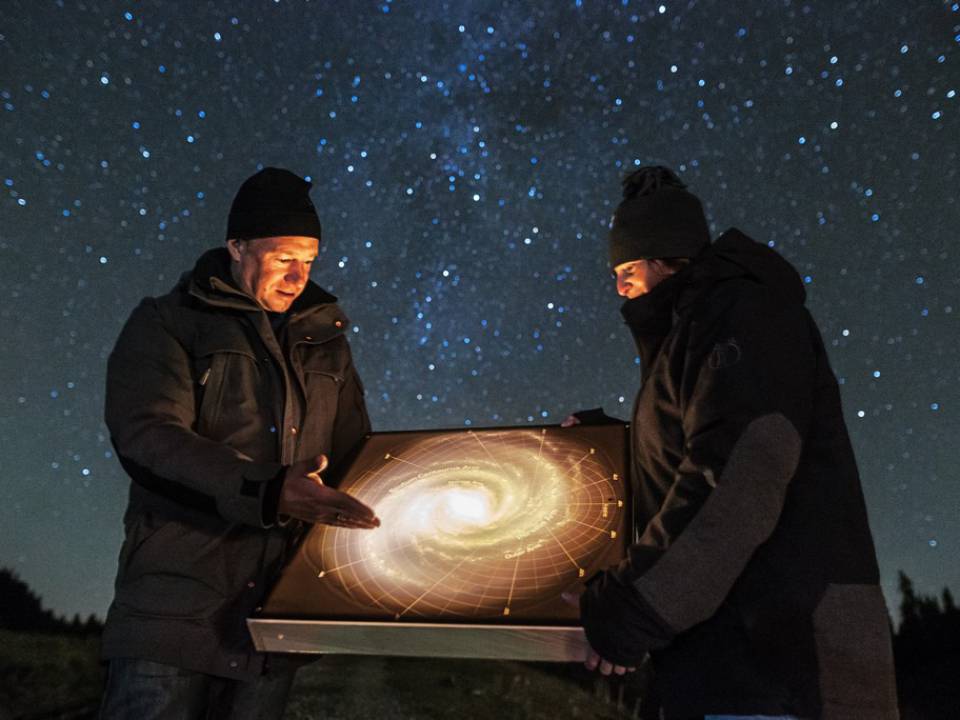 Zwei Männer stehen warm angezogen bei Sternenhimmel draußen und schauen sich ein Sternenbild an