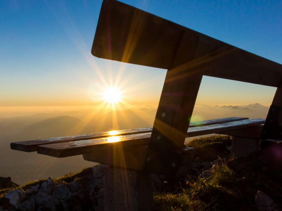 Blick von hinten auf eine Bank und die Sonne, die hinter den Bergen auf geht