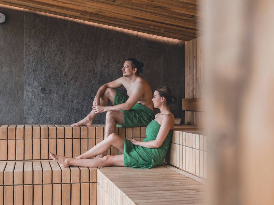 Ein Mann und eine Frau sitzen in der Sauna, tragen grüne Handtücher und genießen ihren Spa-Day im Chiemgau
