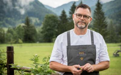 Koch in weißer Kochjacke und grauer Schürze steht vor einem Holzzaun vor einer Wiese