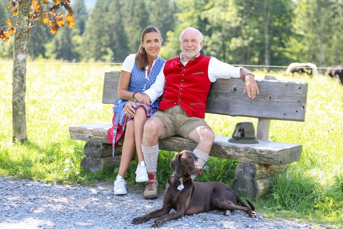 Frau und Herr in Tracht sitzen draußen auf einer Bank, vor ihnen liegt ein Hund