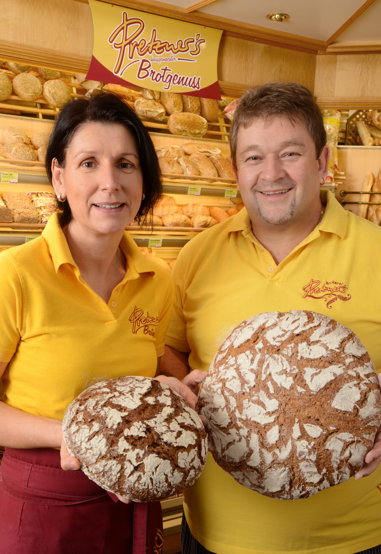 Eine Frau und ein Mann stehen in ihrem Bäckerladen und halten jeweils ein Brot in der Hand