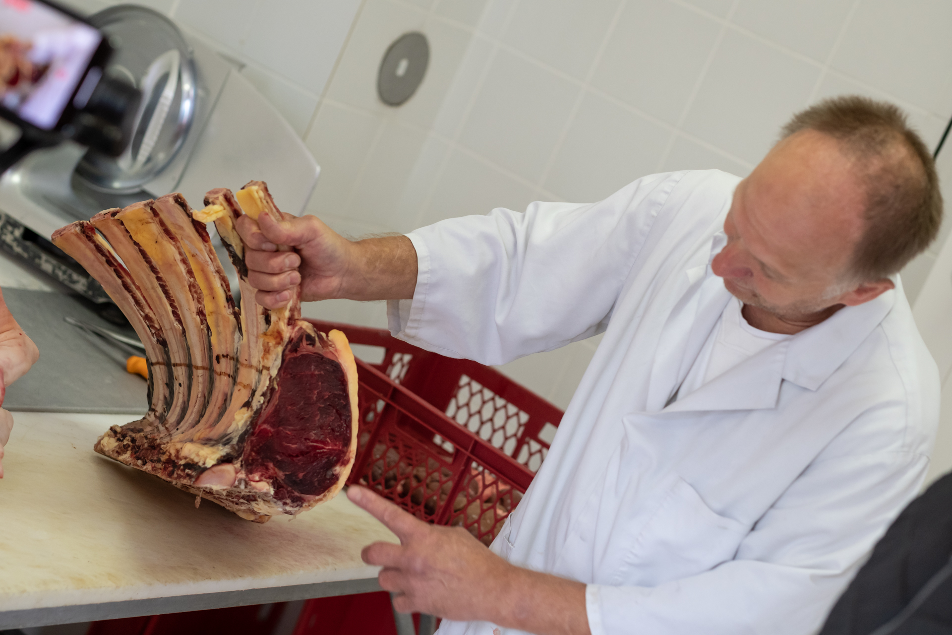 Herr in langer weißer Kochjacke hält ein großes Stück Fleisch mit Knochen und zeigt darauf