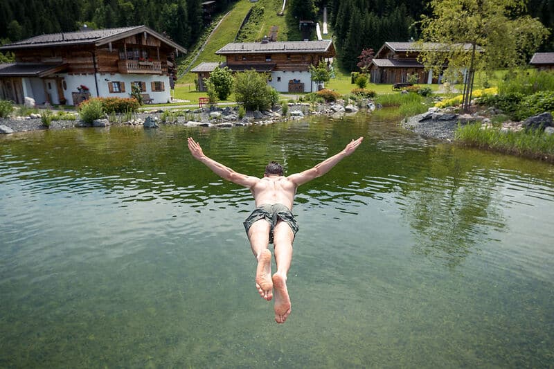 Ein Mann in Badehose taucht mit ausgebreiteten Armen in einen klaren Teich, von hinten gesehen. Umgeben von Holzh&auml;usern, gr&uuml;nem Gras, Felsen und B&auml;umen an einem sonnigen Tag - ein idyllischer Moment in einem Wellnesshotel in Oberbayern.