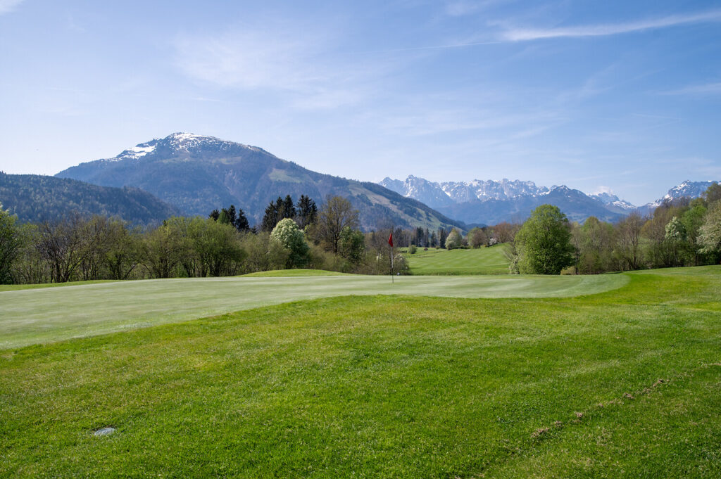 Ein grüner Golfplatz mit einer roten Flagge in der Ferne, umgeben von Bäumen und Bergen mit Schnee auf den Gipfeln unter einem blauen Himmel.