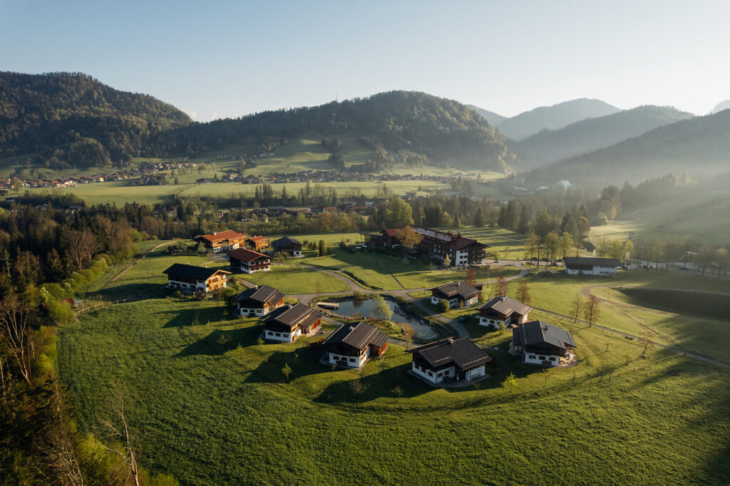 Luftaufnahme eines kleinen Dorfes mit mehreren Häusern, umgeben von grünen Feldern und Hügeln, mit Wäldern und entfernten Bergen unter einem klaren Himmel im frühen Morgenlicht.