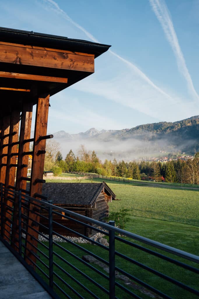 Blick von einem Holzbalkon auf grüne Felder, einen kleinen Holzschuppen, Bäume und ferne Berge, die teilweise von Morgennebel bedeckt sind, unter einem klaren Himmel mit sichtbaren Flugzeugkontrails.