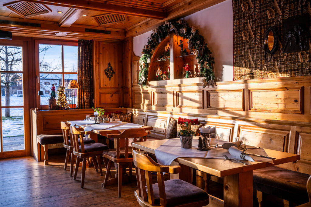Ein gemütliches Holzrestaurant mit Sonnenlicht, das durch ein Fenster fällt, dekoriert mit einem Adventskranz, festlichen Ornamenten und Topfpflanzen auf den Tischen, die zum Essen gedeckt sind.