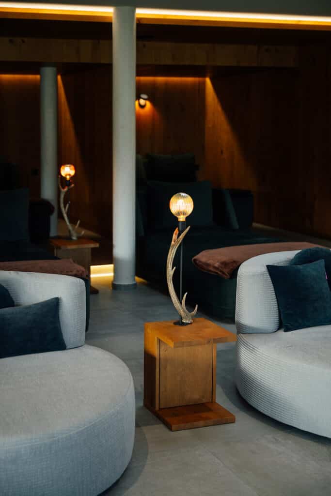 Ein moderner Lounge-Bereich mit runden grauen Sofas, dunklen Kissen und Beistelltischen aus Holz. Auf jedem Tisch steht eine Lampe mit einem geweihförmigen Sockel und einer runden Glühbirne. Die Wände und die Beleuchtung sind warm und gedämpft.