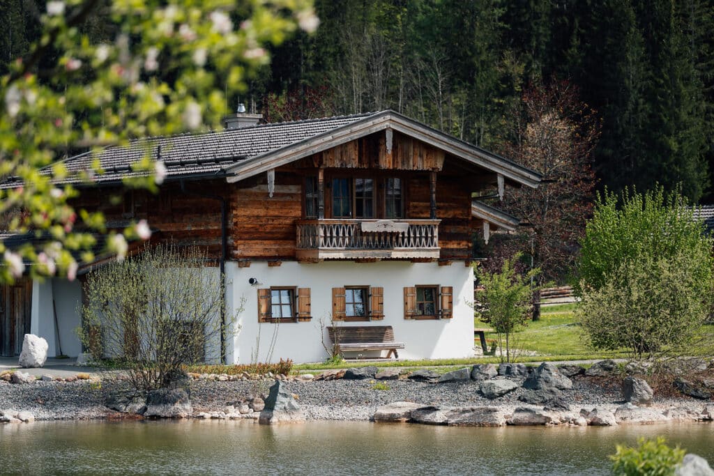 Ein weißes Holzhaus mit Balkon und Fensterläden steht am Rande eines kleinen Teiches, umgeben von Grün und Bäumen im Hintergrund - perfekt für einen ruhigen Aufenthalt in einem Hotel in Reit im Winkl. Vor dem Haus ist eine Holzbank aufgestellt.