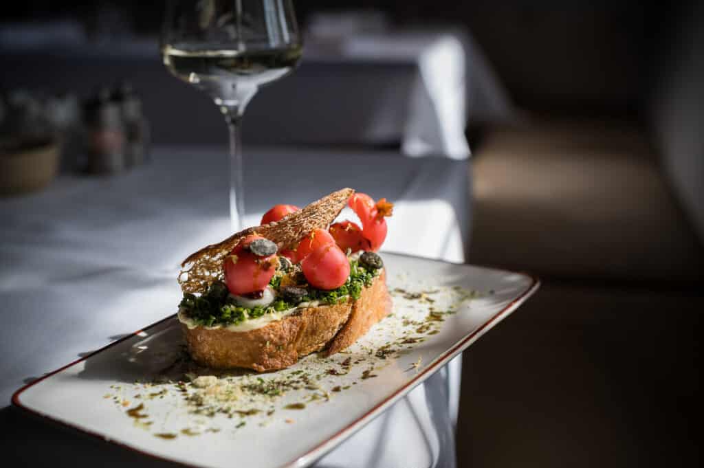Eine Scheibe getoastetes Brot, belegt mit grünem Aufstrich, Kirschtomaten, Sardellen und einem dekorativen Knäckebrot, serviert auf einem rechteckigen Teller mit Kräutern - perfekt für einen Biergartenbesuch in Reit im Winkl, mit einem Glas Weißwein im Hintergrund.