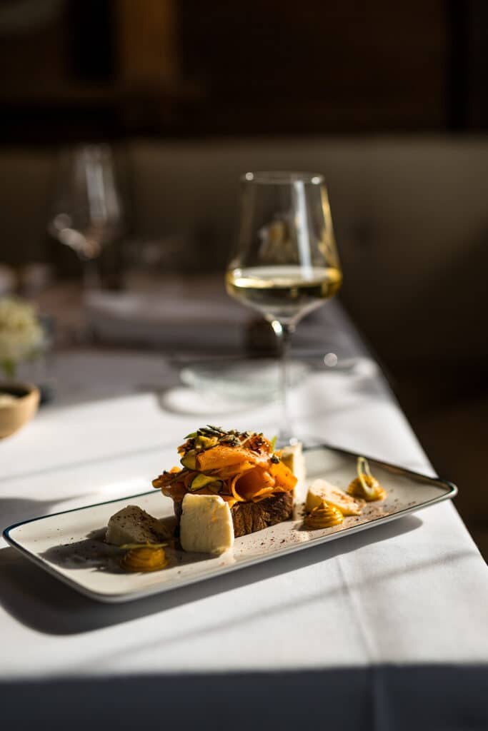 Ein rechteckiger Teller mit einem Dessert, darunter ein Schokoladenkuchen mit Orangenscheiben und Garnitur, serviert mit drei Kugeln Sahne und kleinen Klecksen Soße. Ein Glas Weißwein steht im Hintergrund auf einem weißen Tischtuch.