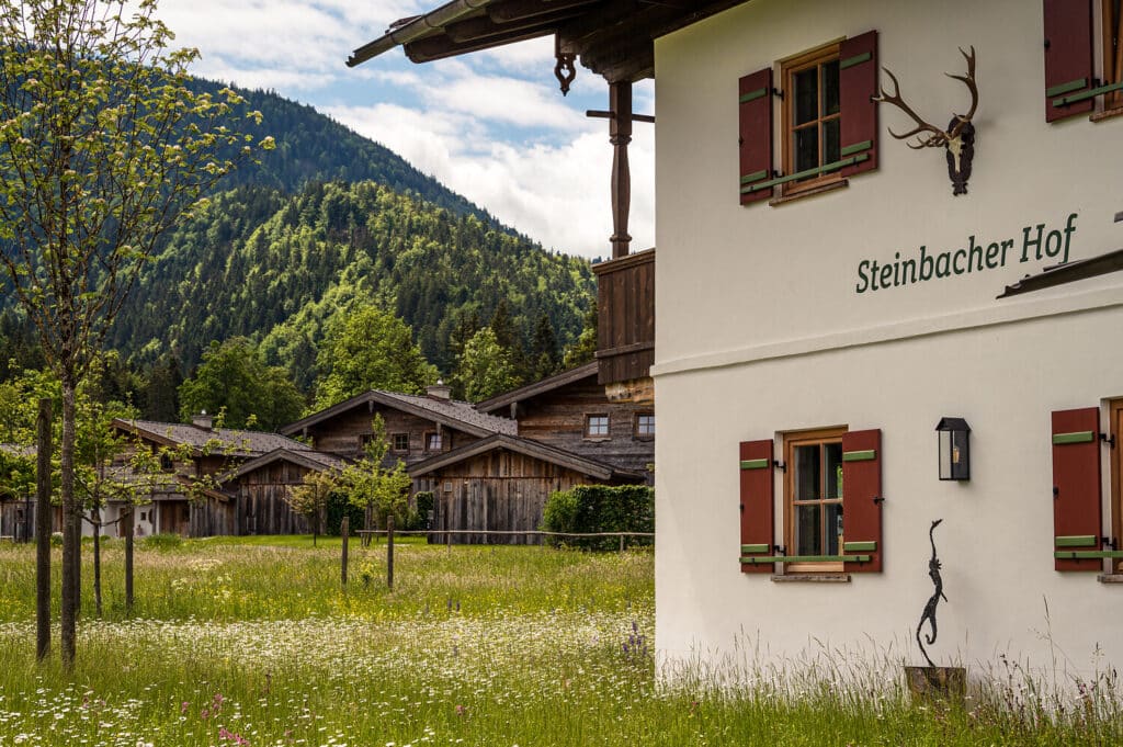 Ein weißes Gebäude mit der Aufschrift "Steinbacher Hof" steht neben einer Wiese mit Wildblumen; dieses charmante Hotel in Reit im Winkl ist umgeben von Holzhütten und bewaldeten Bergen unter einem teilweise bewölkten Himmel.