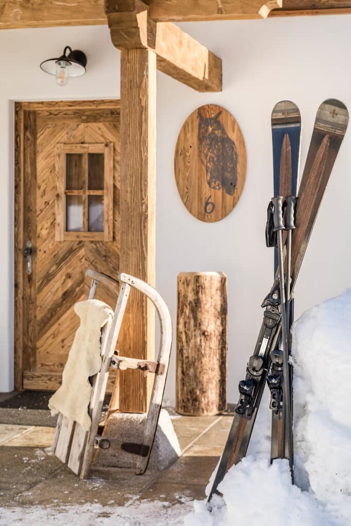 Vor einer rustikalen Holztür stehen ein Paar Holzskier mit Stöcken im Schnee, daneben ein mit einem Schafsfell überzogener Holzschlitten; an der Wand hängen eine Lampe und eine runde Holztafel mit einem Pferdebild.
