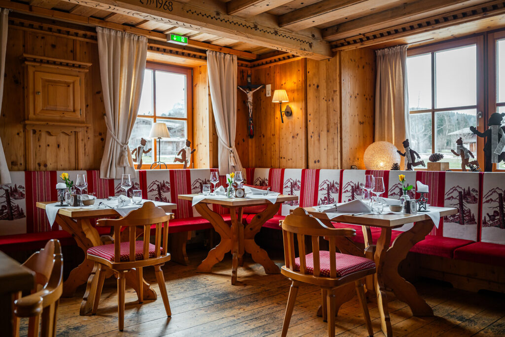 Ein gemütliches Holzrestaurant im Restaurant Heimat, mit rot-weiß gepolsterten Bänken, Holztischen mit Geschirr und Gläsern, Holzstühlen und großen Fenstern, die natürliches Licht hereinlassen. Auf den Fensterbänken stehen dekorative Figuren und eine Lampe.