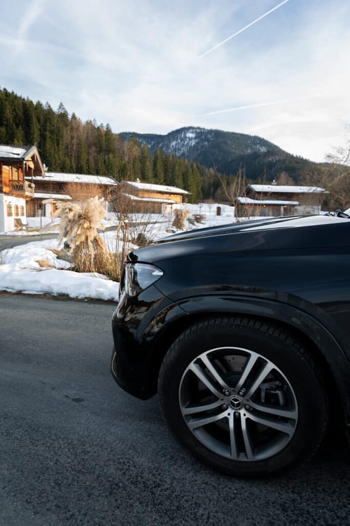 Ein schwarzes Auto, das auf einer Straße geparkt ist, mit verschneitem Boden und Holzhäusern im Hintergrund. Kiefern und Berge sind unter einem teilweise bewölkten Himmel zu sehen.