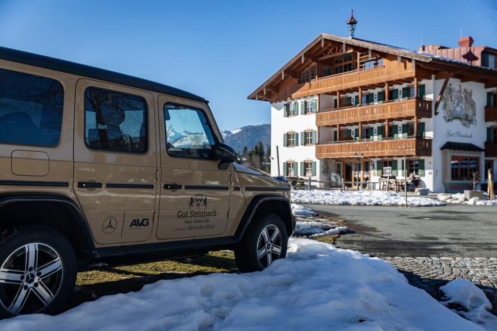 Ein beiger Mercedes-Benz Geländewagen ist an einem klaren, sonnigen Tag auf einem verschneiten Boden neben einem Holzgebäude im alpinen Stil geparkt. Sowohl auf dem Fahrzeug als auch auf dem Gebäude ist das Logo von Gut Steinbach zu sehen.