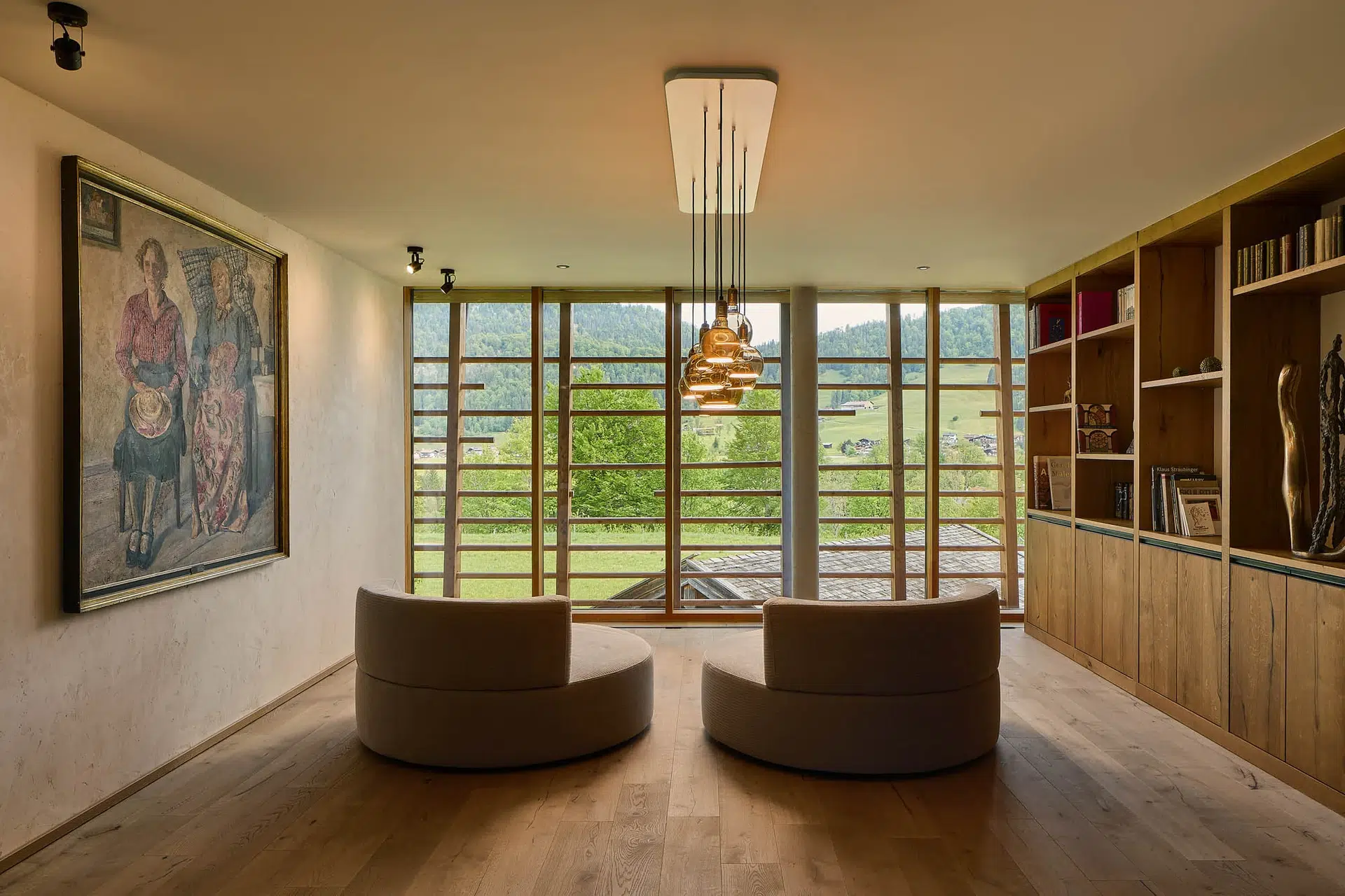 Wellness & Spa 12 Ein modernes Wohnzimmer mit zwei runden beigen Stühlen, Bücherregalen aus Holz auf der rechten Seite, einem großen Gemälde an der linken Wand und raumhohen Fenstern mit Blick auf eine grüne Landschaft. Von der Decke hängen Pendelleuchten.