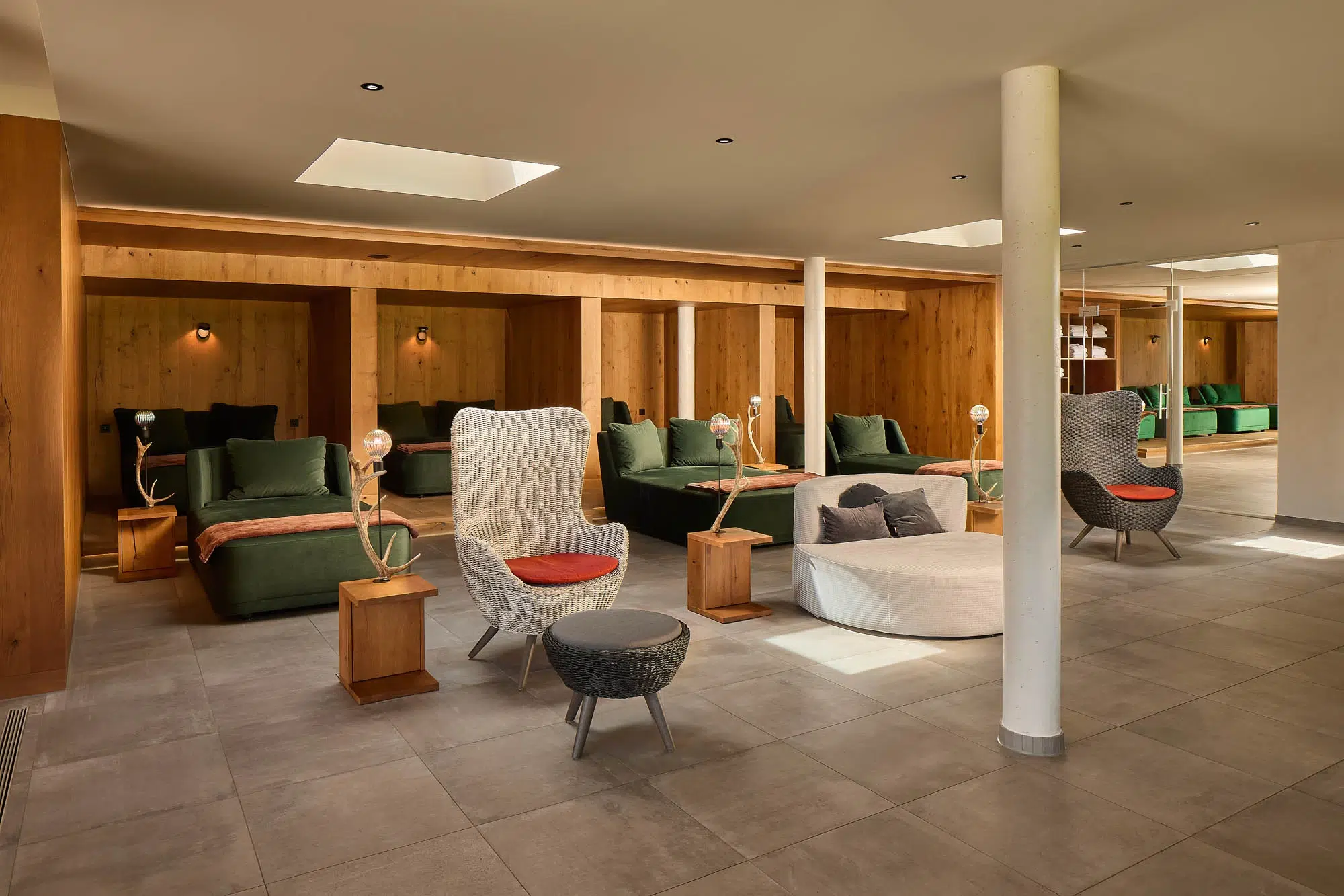 Wellness & Spa 14 Geräumiger Aufenthaltsbereich mit grünen Polsterkissen, Korbstühlen, Beistelltischen aus Holz mit Lampen, Fliesenboden und Holzakzenten an den Wänden; Oberlichter und Deckenleuchten sorgen für die Beleuchtung.