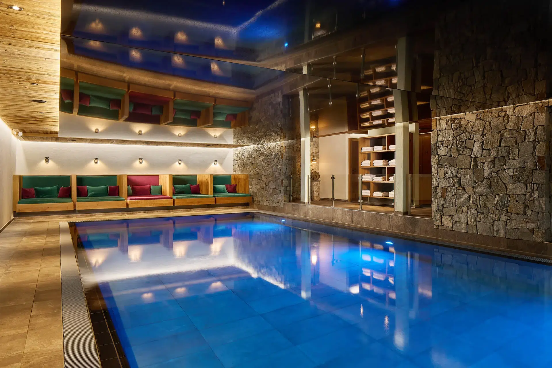 Wellness & Spa 13 Ein Hallenbad in einem modernen Wellnessbereich mit Steinwänden, Holzdecke und eingebauten Sitzgelegenheiten mit bunten Kissen. Im Hintergrund sind Regale mit aufgerollten Handtüchern zu sehen, und Deckenleuchten sorgen für eine warme Atmosphäre.