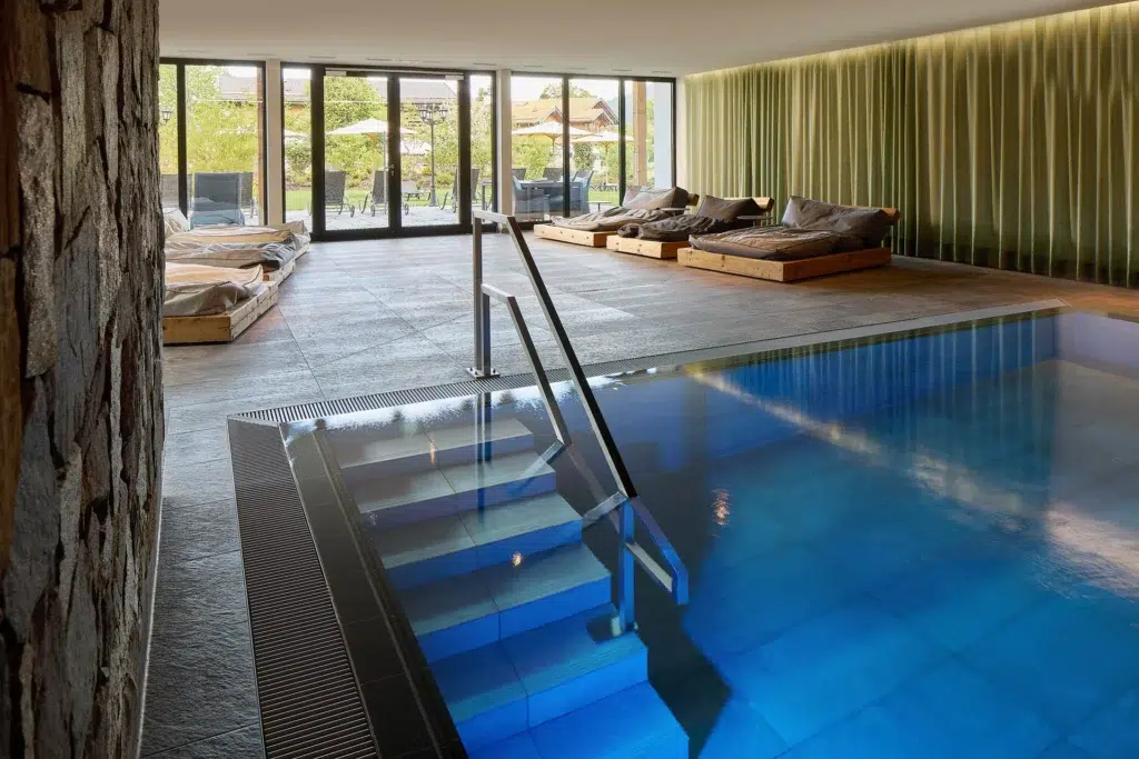 Ein Hallenbadbereich mit klarem, blauem Wasser, einer Steinwand, Fliesenboden und mehreren Holzliegestühlen mit Kissen in der Nähe großer, raumhoher Fenster, durch die Tageslicht hereinströmt. An einer Seite des Raums verlaufen grüne Vertikalpaneele.
