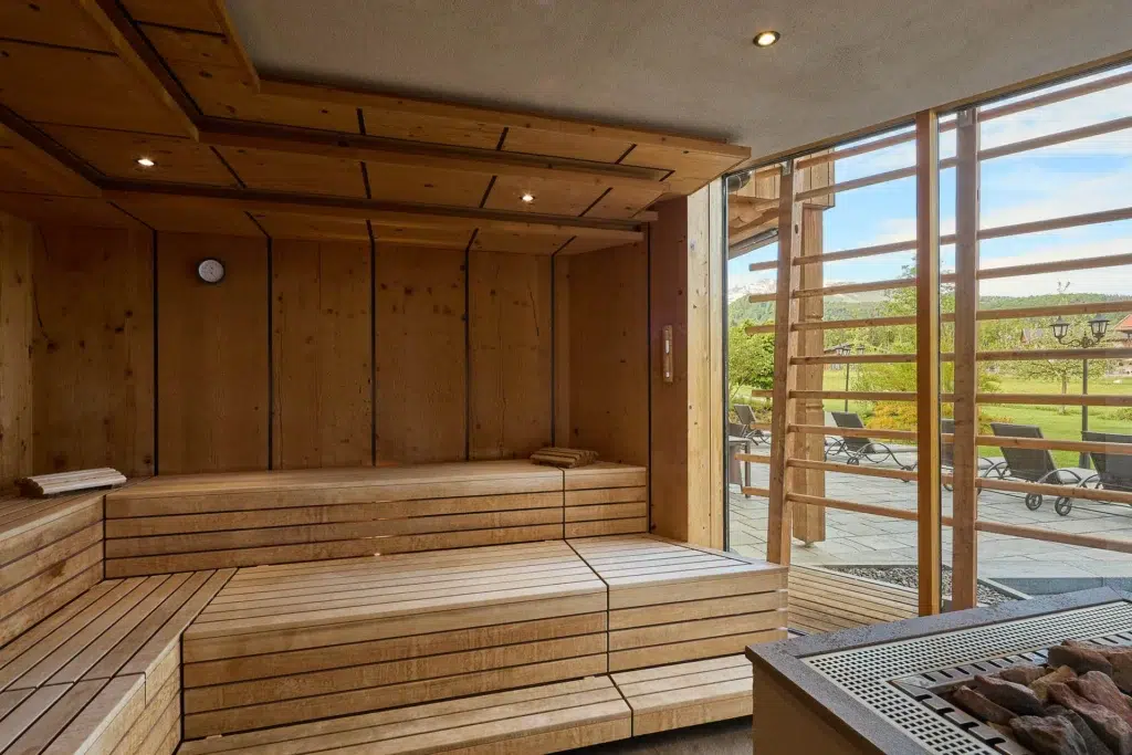 Ein geräumiges Sauna-Interieur aus Holz mit Sitzstufen, gefalteten Handtüchern und einer Uhr an der Wand, neben einem großen Fenster mit Blick auf eine Außenterrasse mit Liegestühlen und viel Grün.