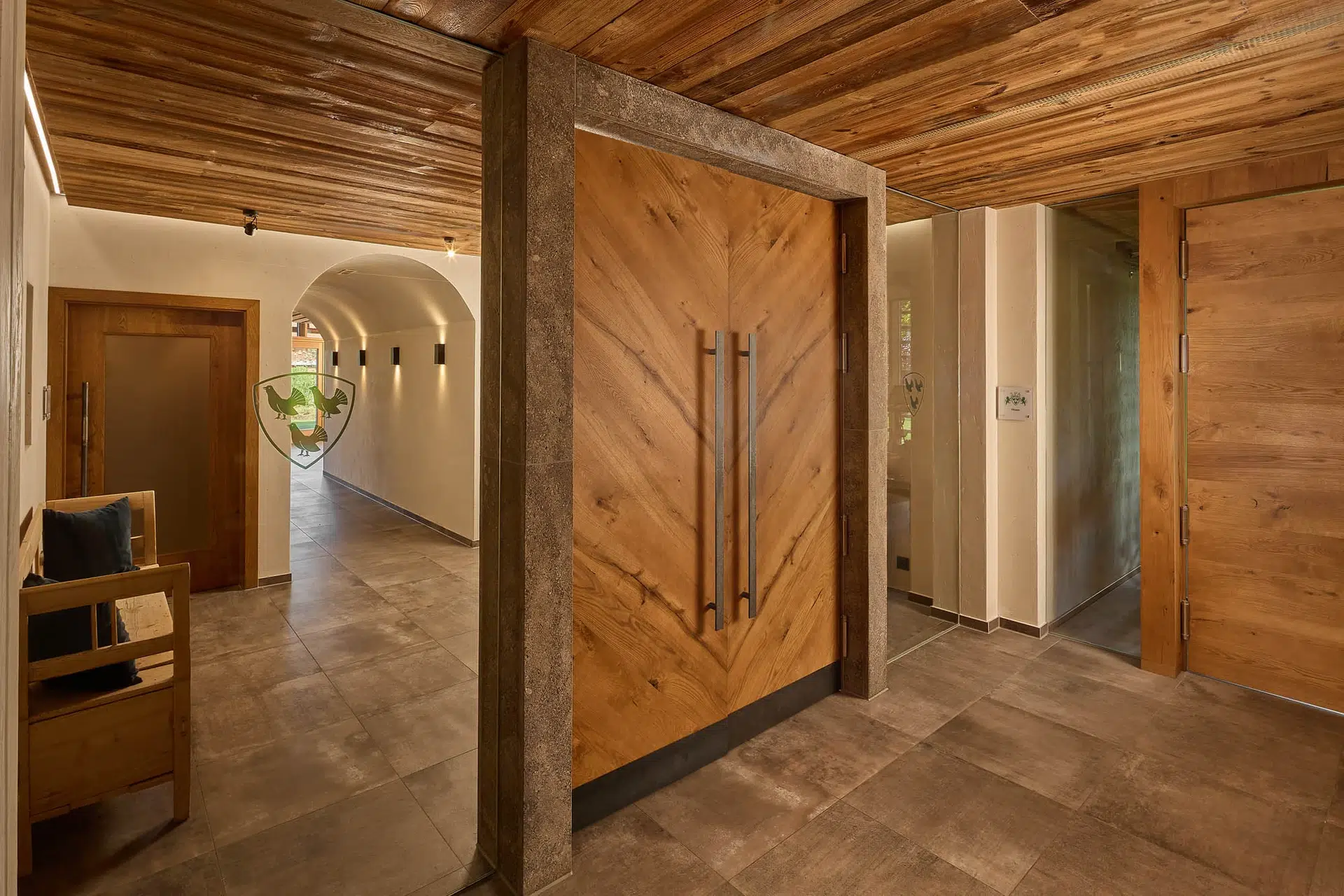 Wellness & Spa 16 Ein moderner Flur mit braunen Fliesenböden, hellen Wänden und einer holzgetäfelten Decke. Große Holztüren mit vertikalen Griffen stehen offen, und an einer Wand steht eine Bank. Von rechts fällt Tageslicht herein.